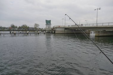 Kappeln 'deki sakin suların üzerine bir balıkçı oltası yerleştiriliyor. Köprü ve arka planda yeşil bir yapı var. Gökyüzü bulutlu, sakin bir atmosfer yaratıyor..