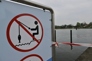 Kappeln 'de balık avlamayı yasaklayan bir tabela. İçinde olta ve kancayla balık tutan birinin resmi var. Arka plan bulutlu bir gökyüzü ile sakin bir su kütlesini gösteriyor..