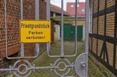 Üzerinde Privatgrundstueck Parken parkının özel mülk olduğunu ve park yasağı olduğunu belirten sarı işareti olan bir giriş kapısı. Arka planda tuğla duvarlar ve bir avlu görüntüsü var..