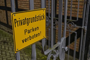 Metal kapıdaki sarı tabela özel mülkü gösteriyor ve park etmeyi yasaklıyor. Arka planda tuğla bir duvar ve işlenmiş demir kapı tasarımı var..