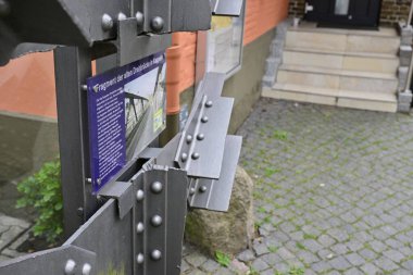 Bir kaldırım taşı yolu ve arka planda bir bina üzerine kurulmuş, mavi bilgi levhası olan metal bir levhanın yakın görüntüsü. Levhada Kappeln 'de sanatsal metal çalışmaları yer alıyor.