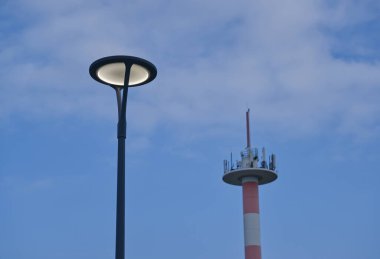 Modern bir sokak lambası bir telekomünikasyon kulesinin arka planında duruyor. Gökyüzü parçalı bulutlu, Kappeln yakınlarındaki Olpenitz 'de sakin bir atmosfer yaratıyor.