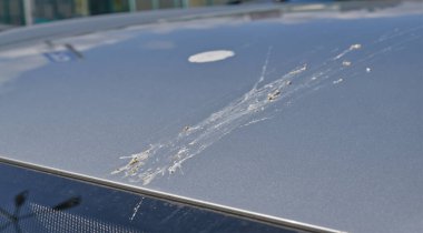 Metalik bir yüzey üzerindeki kuş dışkılarını ve çizgileri gösteren bir arabanın çatı görüntüsü. Güneş ışığı arabadan yansıyor, karmaşayı vurguluyor..