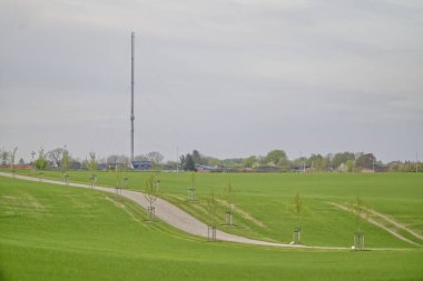 Kollundgaard 'a erişim, tarlaların arasına kurulmuş genç yeşil ağaçlardan oluşan bir caddede uzanan cennet gibi bir yoldur. Arka planda, Kollund radyo kulesi bulutlu bir gökyüzüne karşı.