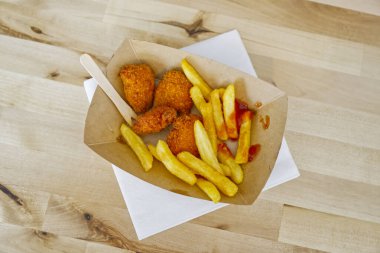 Ekolojik kartondan yapılmış kahverengi bir kâse içinde ahşap çatalla yenmemiş bir patates kızartması. Ayrıca içinde kızarmış tavuk ve ketçap da var..