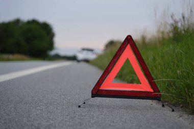 Yolun kenarında bir uyarı üçgeni var, arkasında da arızalı beyaz bir araba.