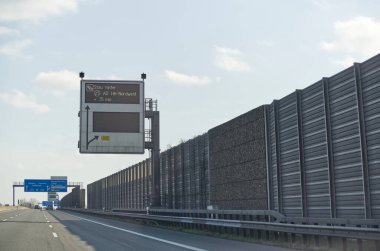 5 dakikalık gecikme uyarısı gösteren dijital trafik işaretinin olduğu bir otoyol sahnesi. Yol, üzerinde açık gökyüzü olan ses bariyerleriyle çevrili..