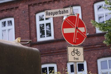 Reeperbahn için sokak tabelası, çapraz çizgili kırmızı dairesel bir dur işareti, ve serbest geçişi gösteren bir bisiklet tabelası. Arka planda pencereli bir tuğla bina görünüyor.