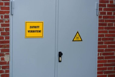 Kırmızı bir binada sarı Zutritt işaretli gri metal kapılar bulunur. Başka bir kapıda ise bir kafatası ve çapraz kemikli üçgen uyarı işareti var..