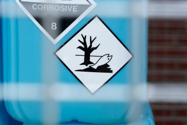 Üzerinde ölü bir ağaç ve balık resmi olan beyaz bir elmas etiketi çevresel tehlike ve doğaya zehirlenmeyi gösteren büyük mavi bir konteynıra iliştirilmiştir.