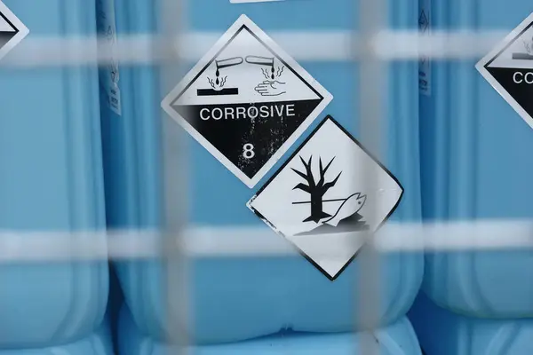 Mavi bir kabın üzerindeki iki tehlike işareti çürüyen bir el ve nesneyle aşındırıcı bir uyarı ve ölü bir ağaç ve ölü bir balığın bulunduğu çevresel tehlike sembolü gösteriyor.