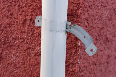 Fotoğrafta yanında metal bir direk olan kırmızı bir duvar var. Direğe eklenmiş kırık bir metal mekanizma, muhtemelen bir mandal veya kilit, hasarlı ve paslı görünüyor.