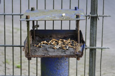 Küçük paslı metal bir kutu tel çitin yanındaki mavi bir direğe bağlıdır. Sigara izmaritleri, kir ve hatta çiğnenmiş bir sakız ile kaplı..