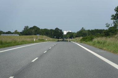 Yeşil tarlalar ve ağaçların arasından geçen iki şeritli bir yol. Uzakta, yol işaretleri görülebilir. Sıcaklık asfaltın üstündeki havayı bulanık gösteriyor..