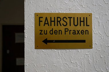 Düz olmayan beyaz duvara dört vidayla asılmış altın renkli tabela sol işaret okuyla Alman metni Fahrstuhl zu den Praxen 'i gösteriyor