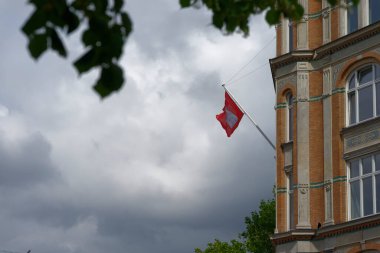 Bulutlu bir gökyüzünün zemininde bir Hamburg bayrağı dalgalanıyor. Mimaride tuğla ve dekoratif elementlerin bir karışımı yer alıyor ve yeşil yapraklar sahneyi kısmen çerçeveliyor..