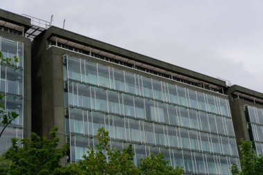 Ön planda büyük cam pencereleri ve yeşilliği olan modern ofis binası bulutlu bir gökyüzüne karşı çağdaş mimari sergiliyor..