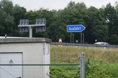 Büyük mavi bir Ausfahrt çıkış tabelasıyla otoyol çıkış yönünü gösteren bir ok ve ön planda beton bina gösteren bir Alman otobanı görüntüsü.