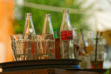 Tahta bir masada iki tepsi boş bardak ve kolalı üç cam şişe. Arka planda, diğer tepsiler ve toplanan çatal bıçaklar görünür..