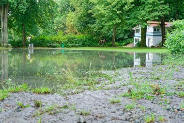 Flensburg Ostseebad 'daki bir halk parkı şiddetli bir yağmur fırtınasından sonra sular altında kaldı. Su çimleri ve yolları kaplıyor, bankları ve oyun alanlarını sular, çevreyi yansıtıyor..