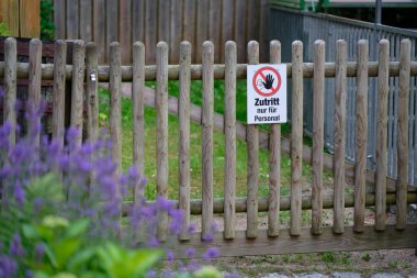 Yeşil bir kırsal alanda küçük ahşap bir kapı duruyor. Almanca bir işaret sadece personele erişimi kısıtlıyor. Yaban çiçekleri ve çimenler yakınlarda yetişir, manzaraya doğal bir bakış verir..