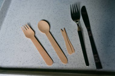 Beyaz tepside tahta bir kaşık, tahta bir çatal, patates kızartması için iki tahta sopa ve metal çatal ve bıçak var. Mutfak aletlerinin karışımı, doğal ve modern yemek aletlerinin bir karışımını gösteriyor..