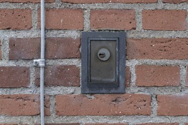 Üzerinde anahtar deliği olan küçük metal bir kutu kırmızı tuğla bir duvara gömülüdür. Kutunun kilitli bir kapısı var ve kullanım amaçlı veya bakım amaçlı gibi görünüyor..