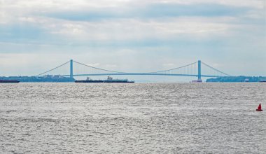 Bir asma köprü, altından geçen gemilerle büyük bir su kütlesi boyunca uzanıyor. Gökyüzü bulutlu ve ön planda kırmızı bir şamandıra görünüyor..