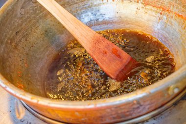 Kavrulmuş bademler için kaynamış kahverengi bir karışım ile dolu derin bir alüminyum kap ateşte duruyor. Tahta bir spatula içinde dinleniyor, kavrulmuş badem hazırlığını öneriyor.