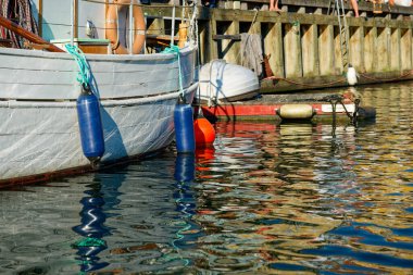 Sessiz bir limanda, çeşitli küçük balıkçı tekneleri kenetlenmiş, her biri büyük şişirilebilir çamurluklarla donatılmıştır. Bu yuvarlak ve uzun tamponlar teknelerin kazınmasını engelliyor.;