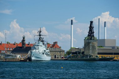 Limanda iki gemi yan yana kenetlendi: beyaz bir yolcu gemisi ve eskimiş görünümlü koyu yeşil bir askeri gemi. Arkalarında, küçük evler ve insanlar hareketli liman sahnesine hayat katıyor..