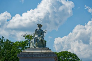 Oturan bir figürün heybetli heykeli, muhtemelen bir tanrıçanın heykeli, taştan bir kaide üzerinde, yemyeşil ağaçlarla çevrili, ve parlak mavi gökyüzü tüylü bulutlarla kaplı..