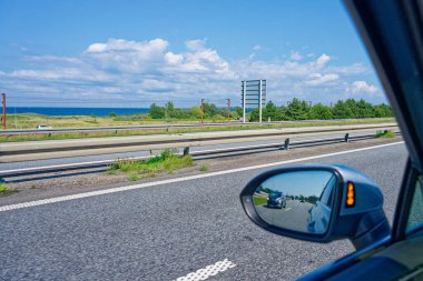 Bir araba yan aynası otobanda başka bir aracı yansıtır. Ayna yanıyor, sola doğru şerit değiştirmeyi öneriyor. Arka planda bariyerler, sakin bir deniz ve parlak mavi bir gökyüzü görülebilir..