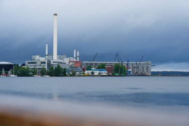 Büyük bir yat limanının yanında suya yakın tesisleri ve boruları olan bir sanayi limanı görülüyor. İnşaat vinçleri yakında yükselirken yağmurlu mavi gökyüzü karamsar bir hava katıyor..
