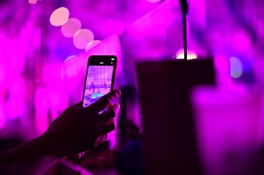 Bir el, canlı neon ışıkla video yakalayan bir akıllı telefonu tutuyor. Telefon ekranı anı yansıtıyor. Teknoloji ve dijital odak yoluyla güzelliğin modern bir sahnesi.