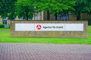 Bir kamu binasında, üzerinde Agentur fuer Arbeit yazan ve Flensburg 'daki istihdam hizmetlerini sembolize eden büyük beyaz bir tabela sergilenmektedir. Arka planda sakin bir şehir ortamını yansıtan ağaçlar görülebilir..