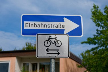 Bir sokak direğinde Alman Einbahnstrasse işareti var. Tek yönlü trafiği gösteriyor. Aşağıda, bir bisiklet tabelası bisikletçilerin iki yöne de gidebileceğini gösteriyor..