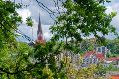 Gotik tarzda sivri bir kilise kulesi Flensburg 'daki yeşil yapraklı bir açıklıktan görülüyor. Aşağıda, renkli iki ve üç katlı küçük bir kasaba katmanlı manzaralı evler yaratıyor..