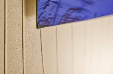 Dikey çizgili bej bir duvar mavi bir kumaşı gevşek bir şekilde asılı tutar. Yatak örtüsüne benzer, basit sahnelere sıradan ve evcil bir dokunuş eklenir..