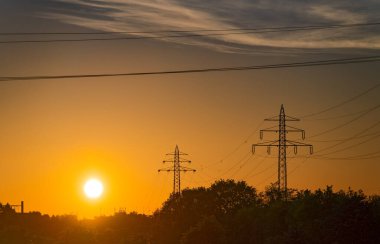 Güneş ufka yaklaştıkça turuncu bir akşam gökyüzü parlıyor. Ağaçların üzerinden yükselen iki elektrik kulesi. Kabloları gün batımını kesiyor. Sakin ve görkemli bir sahne..