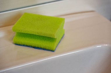 Bir banyo lavabosu açık yeşil bir sünger tutar ve kenarlarında mavi bir ovalama yüzeyi vardır. Banyo ve mutfak yüzeylerini yıkamak ve muhafaza etmek için basit bir temizlik aracı sağlar.