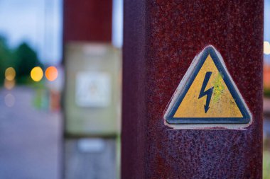 Paslanmış bir metal direk dışarıda duruyor, içinde elektrik tehlikesini gösteren turuncu bir üçgen uyarısı var. Yaşlanmış yüzey elementlere ve uyarılara maruz kalındığını vurgular.