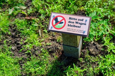 Bir direğe sabitlenmiş bir tabela çapraz ayak işaretli kırmızı bir daire ve Alman metni Bitte auf den Wegen bleiben ziyaretçileri belirlenen yolda kalmaları konusunda uyarır.