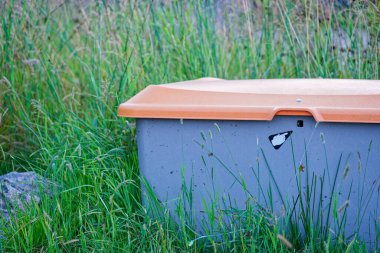 Turuncu kapaklı büyük gri plastik bir kap uzun yeşil çimenlerde duruyor. Kabın üzerindeki bir etiket, kilidi ya da düğmeyi gösteren bir ok içeren beyaz bir el simgesi gösterir.