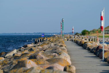 Güneşli bir rıhtım sahnesinde küçük bir patika boyunca büyük kayalar, can simidi direkleri, kırmızı ve yeşil direkler ve deniz kenarında yüzmeye karşı uyarılar görülüyor. Sakin su ışıldıyor.