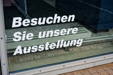 Besuchen Sie unsere Ausstellung adlı cam kapı, camların arkasındaki beyaz ısıtıcı fanı ve çeşitli iç objeleri sergilenen sergi alanına davet ediyor.