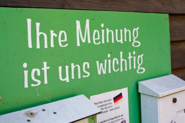 Ahşap bir duvardaki yeşil metal plaka, Almanca ve İngilizce ziyaretçileri bir kağıt formu doldurmaya ve geri bildirim için minnettarlıklarını gösteren beyaz ve açık yeşil posta kutularından birine yerleştirmeye davet ediyor..