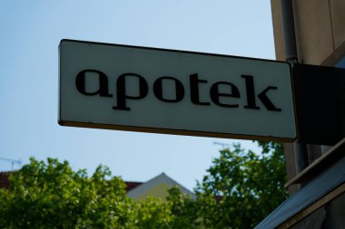 Yüksek bir beton binada kalın siyah harflerle yazılmış Apotek yazan büyük beyaz bir tabela vardır. Arkasında ise Kopenhag CPH kentsel sokak manzarası yer alıyor.