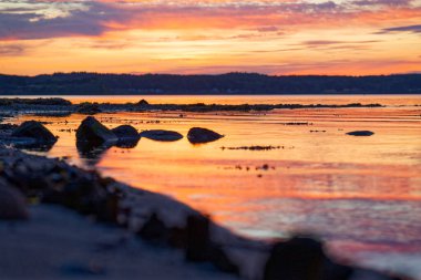 Güneş ufuktan battı ama deniz ve gökyüzü hala turuncu parlıyor. Geniş kayalar sığ sularda dinleniyor ve akşam ışığı yumuşayarak yumuşak bir renge dönüşürken sahil sakinleşiyor..