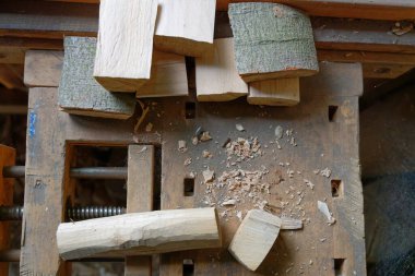 Ahşap bir tezgah, kenarı metal çubuklarla dolu ve el işi için sopa tutan sağlam bir mengene gösteriyor. Yüzeye yayılmış kıvrımlı talaşlarla küçük kesikler ve düzlemsel parçalar etrafa saçılmış..